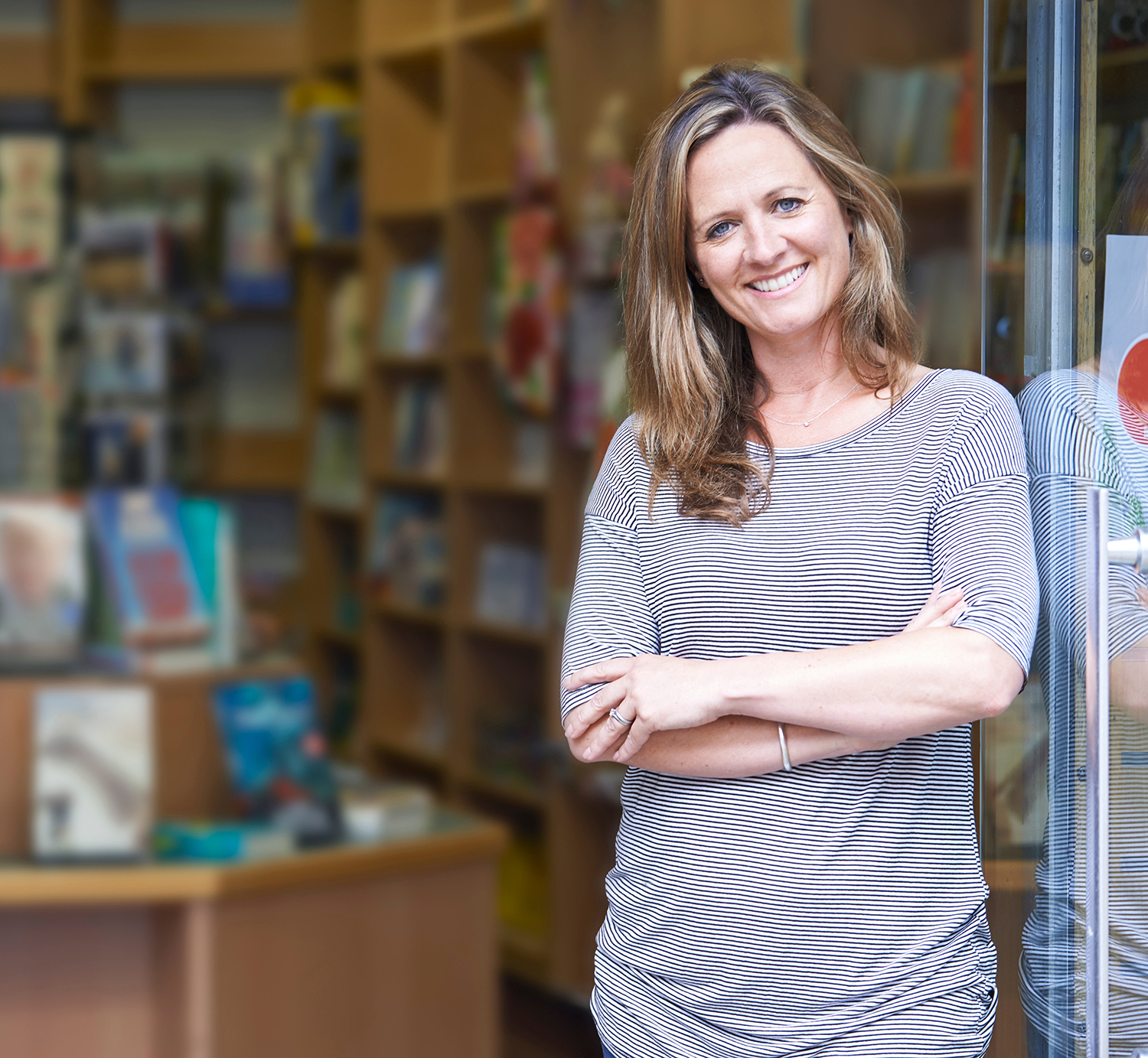 Female shop owner standing in doorway, arms crossed casually in front. Books on shelves in background.
